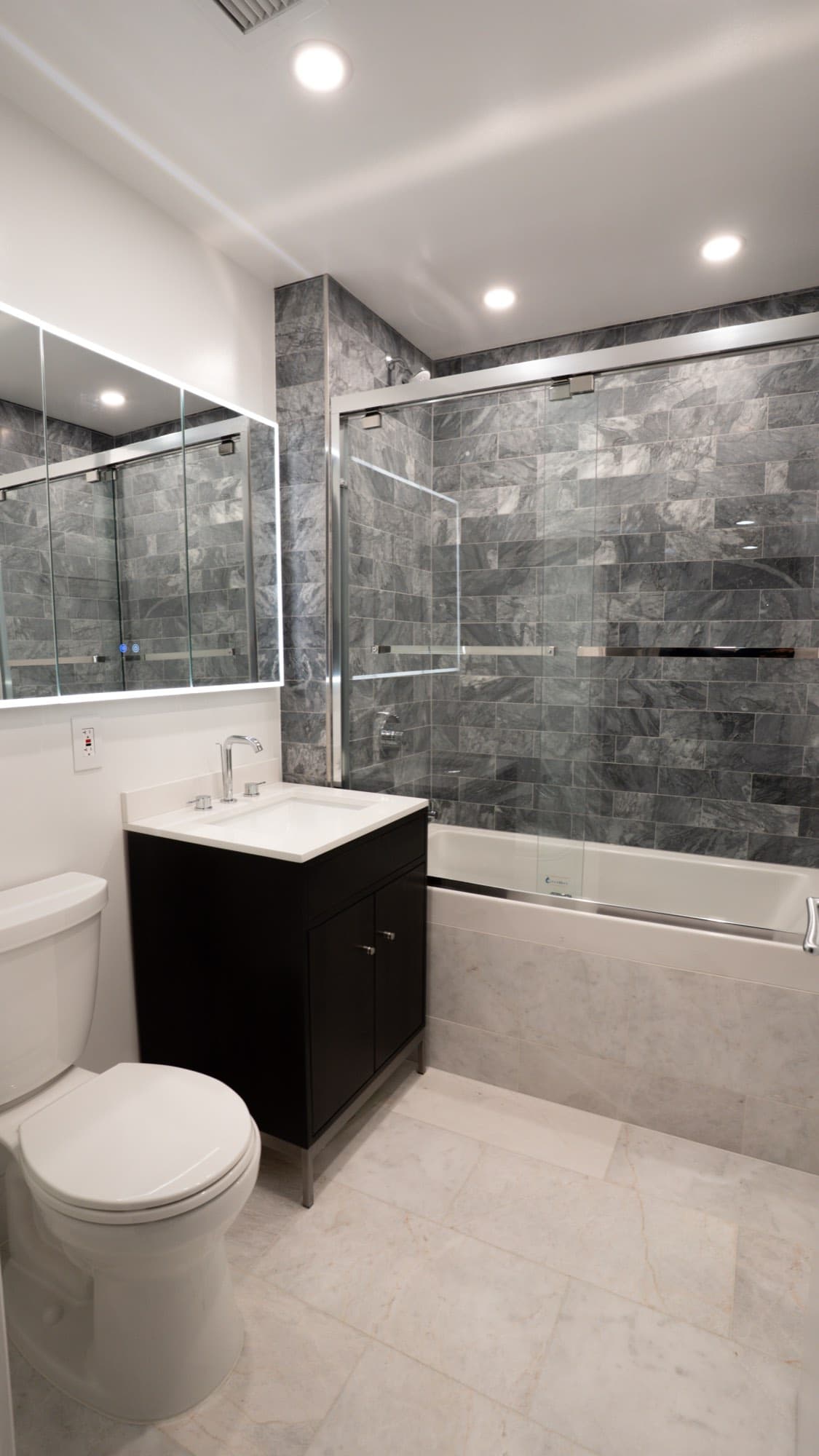 Renovated Dumbo, Brooklyn condo bathroom with gray stone-look tile tub surround, glass enclosure, and backlit mirror